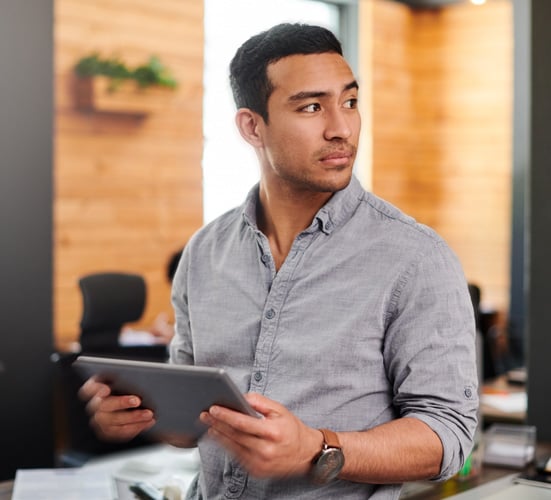 man wearing a grey shirt in the office holding a tablet and looking reflectively to the side